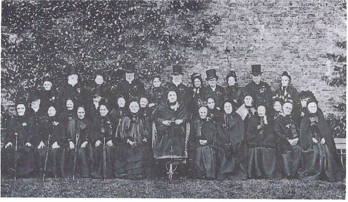 A more formal group of Blue ladies-one enthroned in a wheel chair-in their official gowns and cloaks taken about 1880. The top–hatted gentlemen may be the trustees of the day. The whereabouts of the handsome brick wall can be seen in the picture on page six.